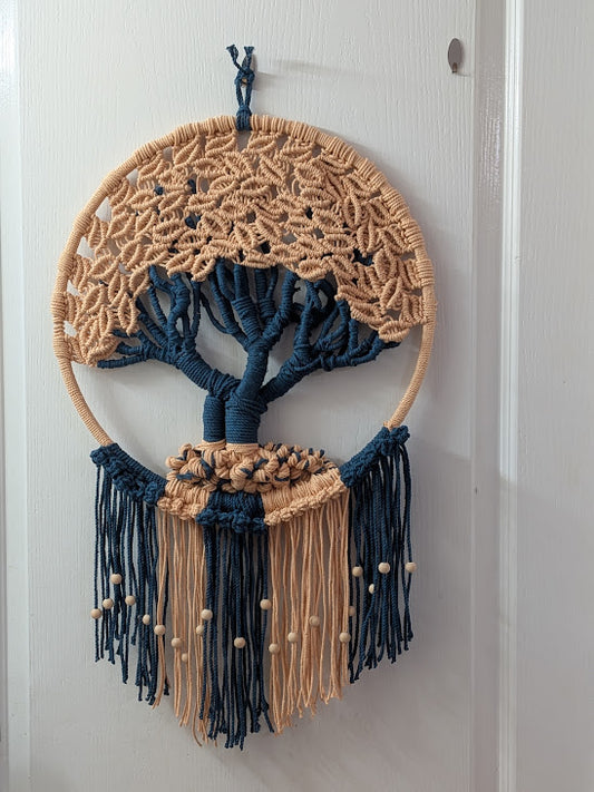 A bohemian macrame wall hanging featuring a tree of life design, suspended from a white door. The circular hoop is made of natural tan cord, with a woven tree design in navy blue and tan, and long fringe tassels hanging below.